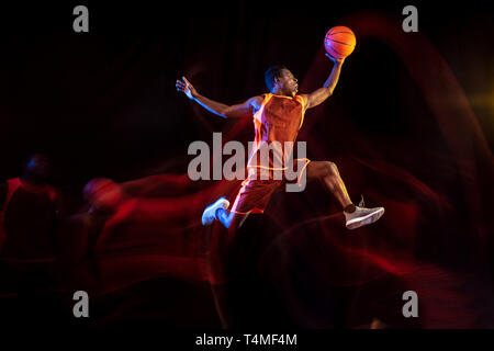 Durst für den Sieg. Afrikanisch-amerikanischen Jungen Basketballspieler der roten Team in Aktion und Neonlicht über dunklen Studio Hintergrund. Konzept für Sport, Bewegung, Energie und Dynamik, gesunde Lebensweise. Stockfoto