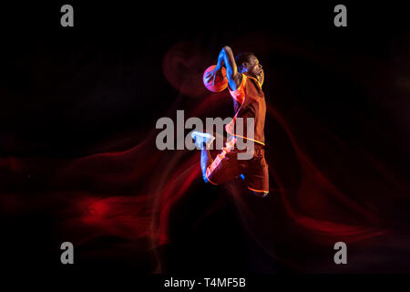 Geheimnisvolle Natur. Afrikanisch-amerikanischen Jungen Basketballspieler der roten Team in Aktion und Neonlicht über dunklen Studio Hintergrund. Konzept für Sport, Bewegung, Energie und Dynamik, gesunde Lebensweise. Stockfoto
