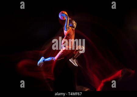 Das Licht von Gewinnen. Afrikanisch-amerikanischen Jungen Basketballspieler der roten Team in Aktion und Neonlicht über dunklen Studio Hintergrund. Konzept für Sport, Bewegung, Energie und Dynamik, gesunde Lebensweise. Stockfoto
