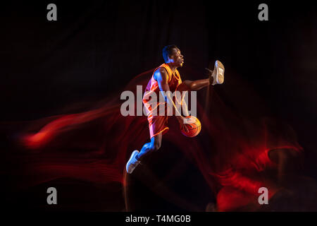 Größer und stärker. Afrikanisch-amerikanischen Jungen Basketballspieler der roten Team in Aktion und Neonlicht über dunklen Studio Hintergrund. Konzept für Sport, Bewegung, Energie und Dynamik, gesunde Lebensweise. Stockfoto