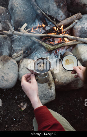 Mann Wanderer gießt sich Kaffee in der Nähe von Feuer. Männliche sitzen und Halten einer Tasse Kaffee nach dem Wandern. Abenteuer Konzept Stockfoto