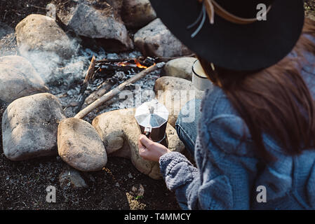 Traveler girl in hat hält heißen Kaffee in einem Wald in der Nähe von Feuer. Weibliche sitzen und Halten einer Tasse Kaffee nach dem Wandern. Trekking-Konzept Stockfoto