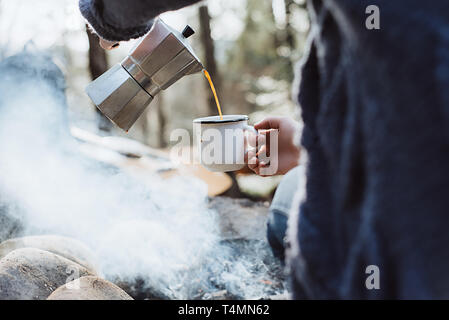 Weibliche Hände Gießen selbst heißen Kaffee in einem Wald in der Nähe von Feuer. Mädchen sitzen und Halten einer Tasse Kaffee nach dem Wandern. Abenteuer Konzept Stockfoto