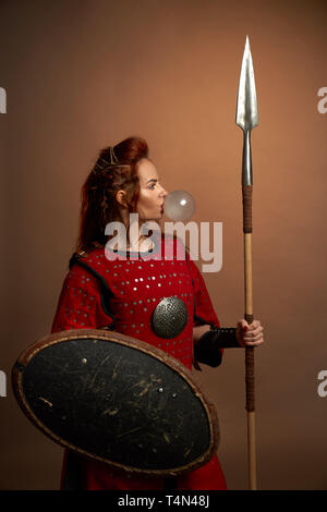 Tapfere weibliche Krieger tragen in Rot mittelalterlichen Kostüm blasen große Blase von Kaugummi. Schön, wunderschöne Frau mit roten Lippen im Studio Posing, Schild und Speer. Stockfoto