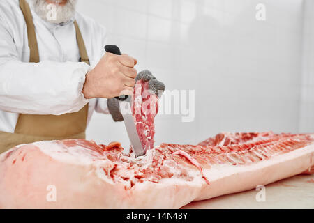 Nahaufnahme von Raw gehacktes Schweinefleisch Schlachtkörper und männlichen Händen mit Messer. Metzger in weißer Uniform und braune Schürze in Fleisch shop arbeiten, Schneiden frisches, rohes Fleisch. Fleisch Fertigung. Stockfoto