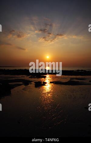 Sunbeam bei Sonnenuntergang casting langen gelben Lichtstrahl über den Ozean. Der Strahl wird in eine Lücke zwischen Steinen und seichten Wasser bei Ebbe wider Stockfoto