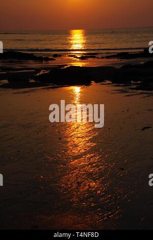 Sunbeam bei Sonnenuntergang casting langen gelben Lichtstrahl über den Ozean. Der Strahl wird in eine Lücke zwischen Steinen und seichten Wasser bei Ebbe wider Stockfoto