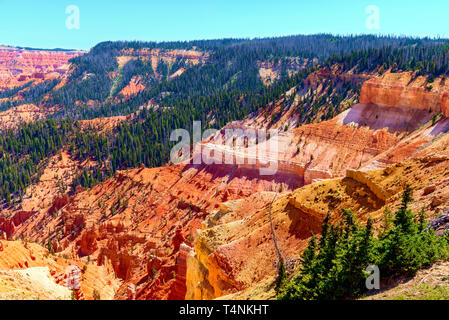 Blick hinunter in eine Schlucht mit grünen Kiefern, gelbe, orange und rote Felsformationen unter einem blauen Himmel. Bunte Canyon Wand mit grünen Bäumen. Stockfoto