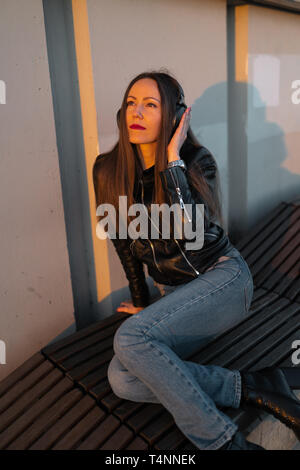 Junge Frau hört Musik in geschlossener Kopfhörer durch ihr Handy tragen eine Lederjacke und Jeans an einem Sonnenuntergang in der Nähe des Flusses Daugava Stockfoto