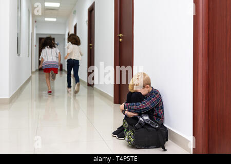 Weinenden kleinen Jungen sitzen in der Schule, mit den Händen auf die Knie und Kopf zwischen seinen Beinen leiden ein Akt von Mobbing während zwei Mädchen laufen weg Stockfoto
