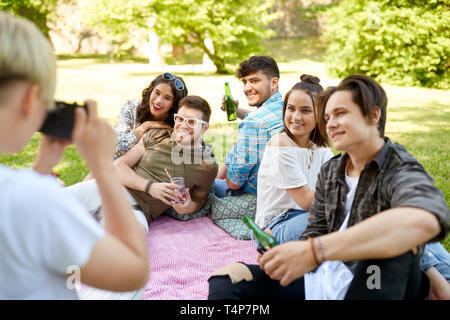 Freundschaft, Freizeitaktivitäten und Technologie Konzept - Frau mit Kamera seine Freunde trinken alkoholfreie Getränke im Sommer Picknick im Park fotografieren Stockfoto