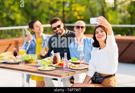 Freizeitaktivitäten und Personen Konzept - glückliche Freunde mit alkoholfreien Getränken, Abendessen auf der Dachterrasse im Sommer und unter selfie per Smartphone Stockfoto
