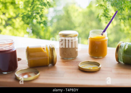 Babynahrung, gesundes Essen und Ernährung Konzept - Gemüse oder Obst pürieren oder Babynahrung in Gläsern und fütterlöffel auf Holzbrett über grüne nat Stockfoto