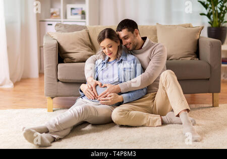 Schwangerschaft, Liebe und Care Concept - glückliche Menschen umarmen schwangere Frau und die Hand Herz Geste zu Hause Stockfoto