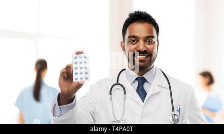 Medizin, Beruf und Gesundheitswesen Konzept - lächelnder Indischer männlicher Arzt im weißen Kittel mit Pillen und Stethoskop über Krankenhaus Hintergrund Stockfoto
