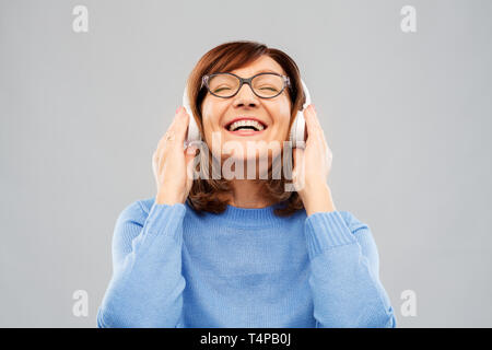 Technik und alte Menschen Konzept - Lächelnde ältere Frau in Brille und Kopfhörer Musik hören über grauer Hintergrund Stockfoto