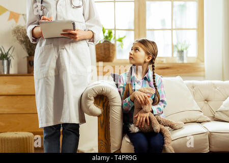 Sorgen über die Diagnose. Kleines süßes braunhaarige Sitzendes Mädchen mit Zöpfen kuscheln Plüsch Spielzeug beunruhigend an einen Kinderarzt mit Noten suchen Stockfoto