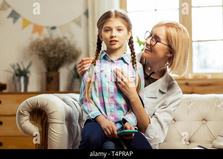 Im Büro des Arztes. Sick hübsches Mädchen mit langen braunen Zöpfe sitzen auf die Knie von angenehmen Lächeln glühende Kinderarzt Cute während Sie exa Stockfoto