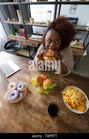 Beeilen Sie sich. Fröhliche Frau am Tisch sitzen und beißen Hamburger Stockfoto