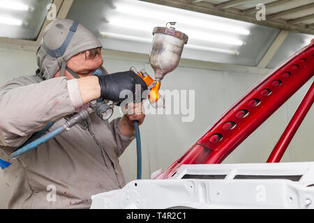 Ein Mann Master in Schutz Overalls und eine Maske hält eine Sprayflasche in der Hand und Sprays rote Farbe auf den Rahmen der Karosserie nach einem Unfall Stockfoto