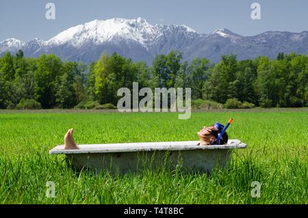 Liegende Frau in einer Badewanne auf einem grünen Feld mit Gras und Bäumen, schneebedeckte Berge, Tessin, Schweiz, Europa Stockfoto