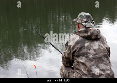 Fischer in Jagd Kleidung sitzen mit einer Angelrute am Ufer. Mann Angeln am See im Frühling, Bäume sind im Wasser reflektiert Stockfoto