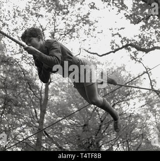 1960, historische, eine junge Abenteuer scout liegend auf einem Seil über seine Hände und Füße sich auf einer Lichtung zwischen zwei Bäumen, England, UK zu manövrieren. Junge Menschen in die Pfadfinder nehmen an vielen Outdoor Aktivitäten, die helfen, ihren Charakter und Leben Fähigkeiten zu entwickeln. Stockfoto