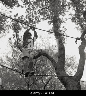 1960 s, historischen, einen Jungen in seine Shorts scout hoch stehende - oben zwischen zwei Seile an einem Baum im Wald, mit Händen und Füßen sich über eine Lichtung, England zu manövrieren, junge Menschen in die Pfadfinder nehmen an vielen Outdoor Aktivitäten, die helfen, ihren Charakter und Leben Fähigkeiten zu entwickeln. Stockfoto