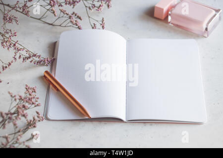 Rosa leere Papier Notebook mit der Goldenen Feder am weißen Tisch mit Blüten und Duft umgeben. Das Konzept von Weiblichkeit Arbeitsplatz Stockfoto