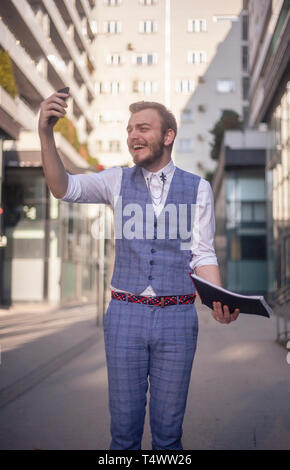 Ein junger Mann, der sein Smartphone in einem Unglauben suchen, im Anzug in einem Geschäftsviertel. holding Notebook in der anderen Hand. Stockfoto