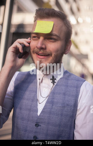 Ein junger Geschäftsmann, Reden auf einem Telefon, mit klebrigen Hinweis auf seine Stirn befestigt. positive Gesichtsausdruck. Stockfoto