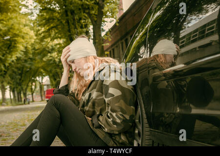 Eine junge Frau mit einer Kopfverletzung lehnt sich gegen ein Auto und sieht schmerzhaft Stockfoto
