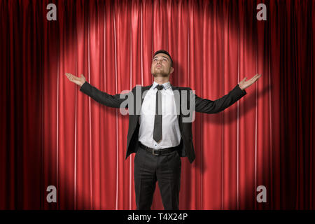 Glückliche junge Unternehmer stehen und sah mit Arme zur Seite ausbreiten von Spotlight beleuchtet, gegen roten Bühnenvorhang. Stockfoto
