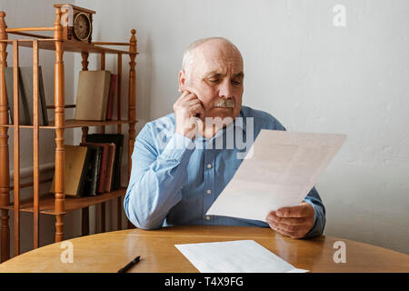 Älterer grauhaariger Mann mit Schnurrbart Lesen eines Handheld Dokument oder einen Brief mit der Hand an seine Wange und einem nachdenklichen ernste Ausdruck sitzt an einem Stockfoto