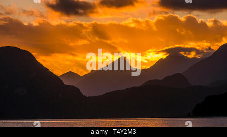 Horizontale schoss der atmosphärischen Golden Sunset piercing Wolken und Bergketten mit Lake Manapouri noch Wasser im Vordergrund von Kepler Track Hütte. Stockfoto