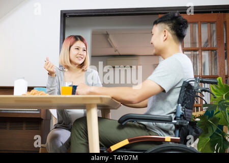 Asiatische paar Frühstück zu Hause. Frau Hand und behinderten Mann im Rollstuhl sitzen am Küchentisch fördern. Paar in Home Konzept Stockfoto