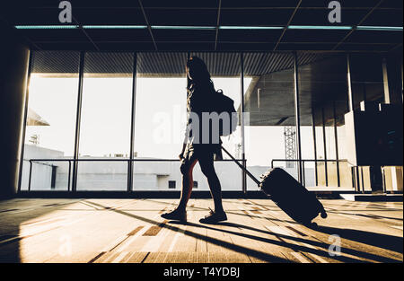 Silhouette von jungen Mädchen zu Fuß mit Gepäck zu Fuß am Flughafen-terminal-Fenster in der Sonnenaufgangszeit, Reisekonzept, Reise-Lebensstil. Stockfoto