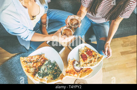 Junge Frauen jubeln Bier und essen Pizza in ihrer Wohnung - zwei Mädchen mit einem Mittagessen gemeinsam mit hausgemachten Speisen Stockfoto