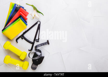 Ansicht von oben Bild von Kurzhanteln, bunte elastische Gummi Expander, Bremssättel und Maßband auf einem weißen strukturierten Hintergrund. Gesunder Lebensstil, Fitnessraum und nicht Stockfoto