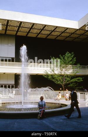 Ein männlicher Fußgängerzone betritt den Rahmen, als lächelnde Frau, sitzt auf der Kante einer Runde Brunnen, posiert vor einer Ausstellung Pavillon, in New York World's Fair, Flushing Meadows Park, Queens, New York, Juni 1964. () Stockfoto