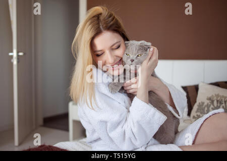 Schöne blonde junge Mädchen in einem Terry White Robe in ihrer Wohnung umarmt sie geliebte Katze. charmante junge Frau und ihr Haustier grauen lop-eared cat Stockfoto