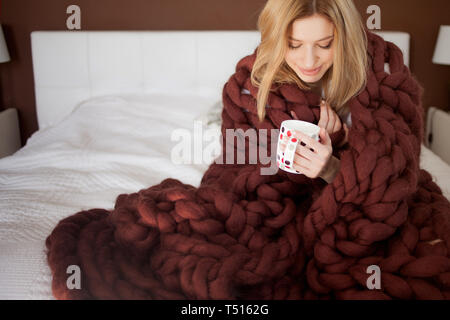 Nette junge Frau sitzt auf dem Bett in einem großen und flauschige Brown plaid gewickelt. Schöne Mädchen in einer gemütlichen Decke mit einer Tasse Tee. Wärme und Komfort Stockfoto