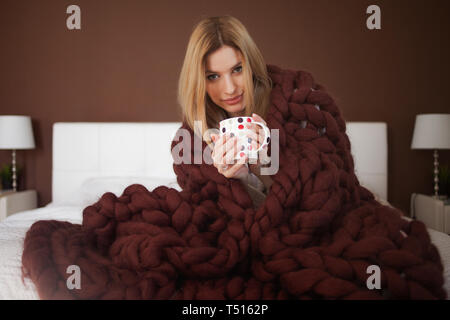 Nette junge Frau sitzt auf dem Bett in einem großen und flauschige Brown plaid gewickelt. Schöne Mädchen in einer gemütlichen Decke mit einer Tasse Tee. Wärme und Komfort Stockfoto