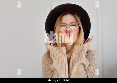 Charmante junge Frau in einem weicher Pullover und Gläsern, die in einem schwarzen Hut gefühlt. Porträt einer Glücklich lächelnde Mädchen sendet einen Kuss Stockfoto