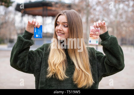 Die junge Dame zeigt Plastikkarte und Cash zu Kamera beim Stehen in Park Stockfoto