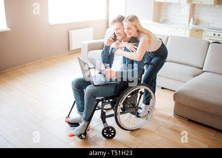 Junge Menschen mit besonderen Bedürfnissen. Das Sitzen auf Rollstuhl und am Telefon sprechen. Junge Frau ihn umarmen. Stehend von hinten. Laptop auf den Knien Stockfoto
