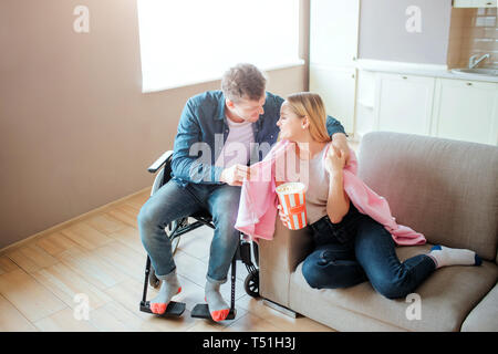 Junger Mann mit Einbeziehung der Freundin. Er umfasst die Schulter Decke und Lächeln. Mit besonderen Bedürfnissen Person. Sitzung am Rollstuhl. Stockfoto