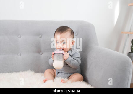 Kleinen niedlichen Baby Mädchen sitzen im Zimmer auf dem Sofa das Trinken der Milch aus der Flasche und lächelnd. Glückliches Kind. Familie Menschen indoor Raumkonzepte. Kindheit b Stockfoto