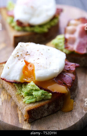 Pochiertes Ei Toasts mit Avocado und Speck. Auf einem Holzbrett serviert, hohe Auflösung Stockfoto