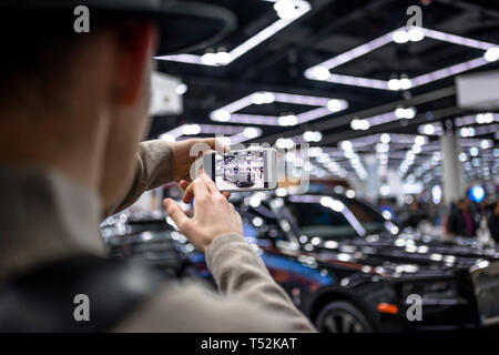 Ein Mann in einem Hut ist das Fotografieren auf einem Handy ein schwarzes Auto Crossover glitzernden funkeln wie er, in der Lobby stehend in der Belichtung hangin Stockfoto